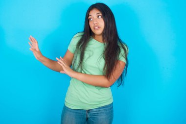 Mavi arka planda yeşil tişört giyen genç İspanyol kız ellerini boş alana uzatıyor ve yakına gelmemesini söylüyor.