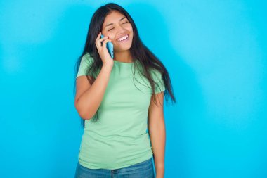 Mavi arka planda yeşil tişört giyen komik İspanyol kız mutlu bir şekilde gülüyor, telefon görüşmesi yapıyor, arkadaşı tarafından eğlendiriliyor, gözlerini kapatıyor..