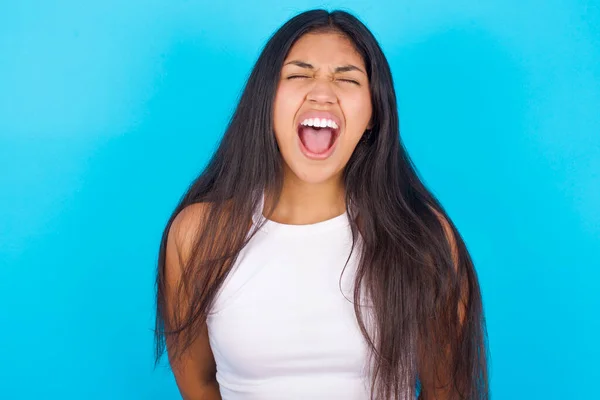 Stressful Young hispanic girl wearing white tank top over blue background  screams in panic, closes eyes in terror, keeps hands on head, finds out terrified news, can\'t believe it.