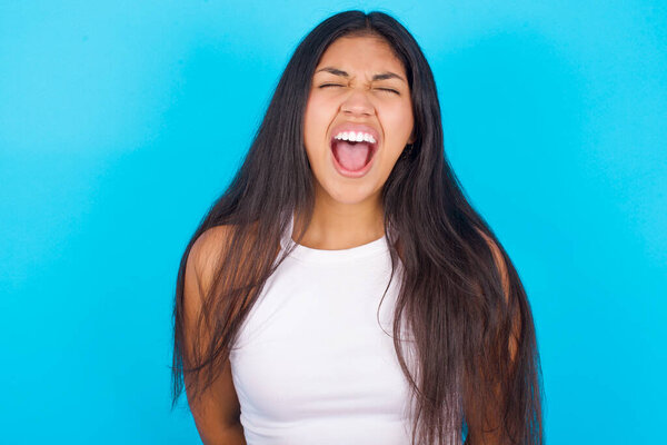 Stressful Young hispanic girl wearing white tank top over blue background  screams in panic, closes eyes in terror, keeps hands on head, finds out terrified news, can't believe it.