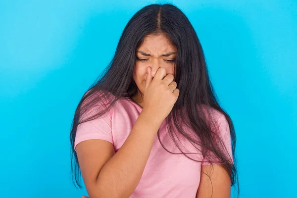 青い背景にピンクのTシャツを着た若いヒスパニック系の女の子は 悪臭のために彼の鼻を保持 — ストック写真