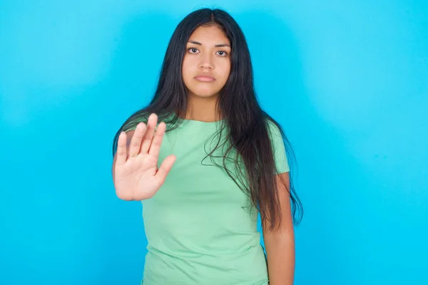 青い背景の上に緑のTシャツを着た若いヒスパニック系の女の子は ストップサイン禁止シンボルは 厳格な表現でカメラに手のひらを前方に保つ示しています — ストック写真