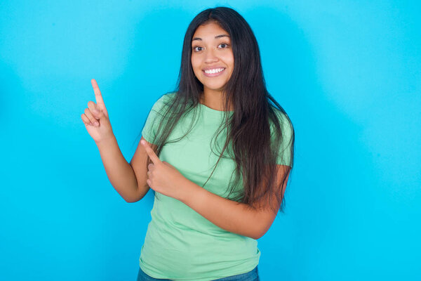 Young hispanic girl wearing green T-shirt over blue background  points at copy space indicates for advertising gives right direction