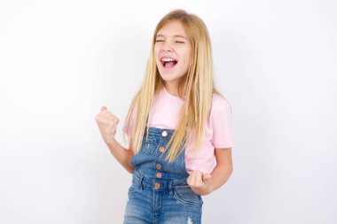 Beyaz arka planda kot pantolon giyen güzel beyaz bir kız çok mutlu ve heyecanlı bir şekilde kollarını kaldırıp, gülümseyerek ve başarı için çığlık atarak kazanan jestler yapıyor. Kutlama konsepti.