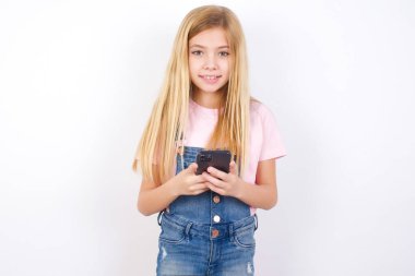 Beyaz arka planda kot pantolon giyen güzel beyaz kız uzak iletişimden zevk alıyor, cep telefonu kullanıyor, hızlı ve sınırsız internet sörfü yapıyor, hoş bir gülümsemesi var, online alışveriş yapıyor.,