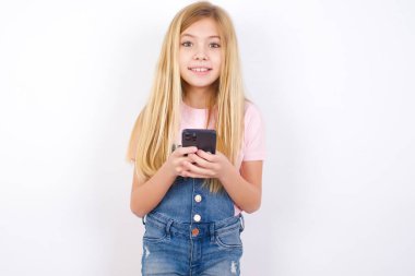 Heyecanlı, beyaz arka planda kot pantolon giyen güzel beyaz bir kız. Elinde akıllı telefon var ve iyi haberler aldıktan sonra kameraya hayret içinde bakıyor..