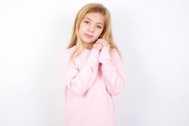 Beyaz arka planda pembe süveter giyen güzel beyaz bir kız ellerini kameraya doğru nazikçe gülümsüyor.