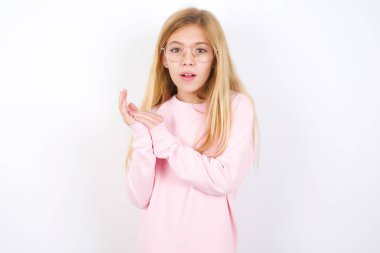 Şaşırmış duygusal, güzel, beyaz arka planda pembe kazak giyen beyaz kız avuçlarını ovuşturur ve kameraya inançsızlıkla bakar.