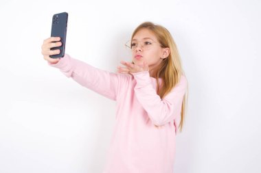 Beyaz arka planda pembe kazak giyen güzel beyaz bir kız akıllı telefon kamerasına hava öpücüğü verip selfie çekiyor ve internetten Mwah 'ı gönderiyor..