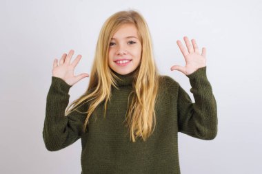 Beyaz arka planda yeşil örülü süveter giyen beyaz çocuk on numarayı gösterirken kendinden emin ve mutlu bir şekilde gülümsüyor..