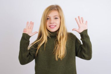 Beyaz arka planda yeşil örülü süveter giyen beyaz çocuk dokuz numaralı parmaklarını göstererek gülümsüyor ve mutlu..