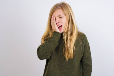 Beyaz arka planda yeşil örgü süveter giyen beyaz çocuk esnemekten yoruldu. Yüzün yarısını, gözünü ve ağzını eliyle kapattı. Yüzüm acıyor..