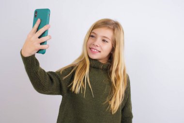 Beyaz arka planda yeşil örgü süveter giyen beyaz bir kız gülümsüyor ve sosyal medyasında paylaşmaya hazır bir selfie çekiyor..