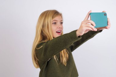 Beyaz arka planda yeşil örülü süveter giyen beyaz bir kız sosyal medyaya göndermek için selfie çekiyor ya da arkadaşlarıyla görüntülü görüşme yapıyor..