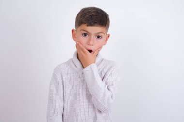 Beyaz arka planda dikilen örgü süveter giyen duygusal beyaz çocuk şaşkınlıktan soluk soluğa kalmış, ağzı palmiyeyle kapatılmış, kameraya şok olmuş gibi görünüyor. Tepki konsepti