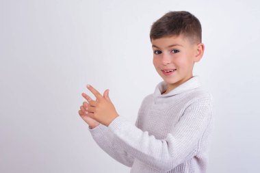 Beyaz arka planda dikilen örgü süveter giyen beyaz çocuk, en düşük fiyatları gösteren boş bir parmak gösteriyor, kameraya bakıyor.