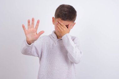 Beyaz arka planda dikilen örgü süveter giyen beyaz çocuk avuçla gözlerini kapar ve dur hareketi yapar, herkesten saklanmaya çalışır..