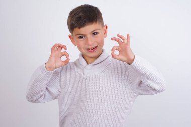 Beyaz arka planda dikilen örgü süveter giyen beyaz çocuk iki elini de OK işaretinde gösteriyor. Onaylama veya öneri kavramı
