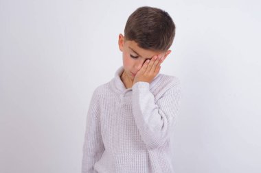 Beyaz arka planda dikilen örülü süveter giyen yorgun beyaz çocuk uykulu bir ifadeye, kasvetli bir bakışa sahip, yüzü elle kaplanmış, gözleri kapalı, yorgunluktan nefesi kesilmiş, partiden sonra bitkinlik ve bitkinliğe sahip.
