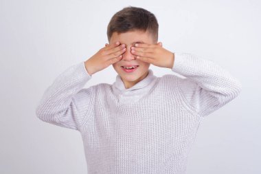 Beyaz arka planda dikilmiş örgü kazak giyen beyaz çocuk elleri gülücüklü ve komik bir şekilde gözlerini kapatıyor. Kör kavram.