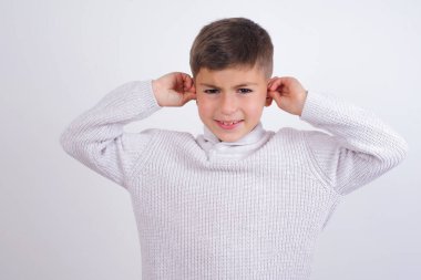 Beyaz arka planda dikilen örgü kazak giyen beyaz çocuk yüksek sesli müzik sesi için kulaklarını parmaklarıyla kapatıyor. Sağır kavramı.