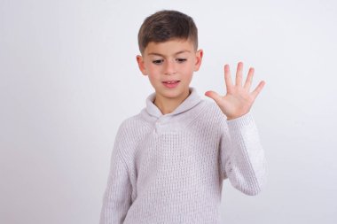 Beyaz arka planda duran örgü süveter giyen beyaz çocuk beş numarayı gösterirken kendine güvenen ve mutlu bir şekilde gülümsüyor..