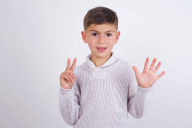 Beyaz arka planda duran örgü süveter giyen beyaz çocuk yedinci parmaklarını gösterip gülümseyerek mutlu ve kendinden emin görünüyor..