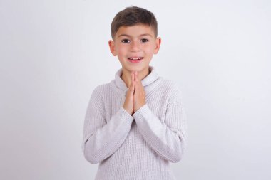 Beyaz arka planda örülü süveter giyen beyaz çocuk elleriyle dua edip af diliyor kendinden emin bir şekilde gülümsüyor..