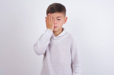 Beyaz arka planda duran örgü süveter giyen beyaz çocuk esnemekten yoruldu. Yüzün yarısını, gözünü ve ağzını eliyle kapattı. Yüzüm acıyor..