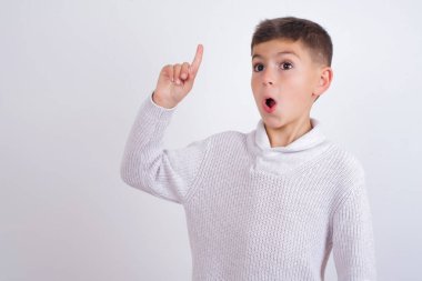 Beyaz arka planda dikilmiş örgü süveter giyen beyaz çocuk. Parmakla işaret ediyor ve dahiyane düşüncelerden esinlenerek bakıyor. İyi fikir işareti gösteriyor.