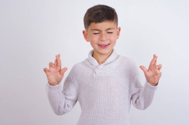 Beyaz arka planda dikilen örgü süveter giyen beyaz çocuk umut ve gözler kapalı gülümseyerek el kol hareketi yapıyor. Şans ve batıl inanç..