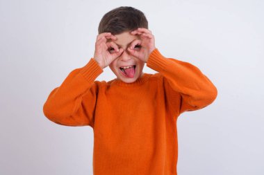 Beyaz arka planda dikilmiş örgü turuncu süveter giyen beyaz çocuk dürbün gibi jest yapıyor, gözleri parmaklarından bakıyor. Çılgın ifade.