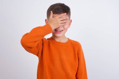 Beyaz arka planda dikilmiş örgü turuncu süveter giyen beyaz çocuk. Gülümsüyor ve elleri yüzünde şaşkınlıktan gözleri kapayarak gülüyor. Kör kavram.
