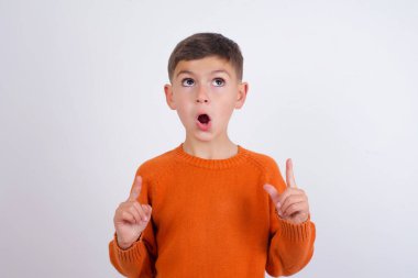 Beyaz arka plan üzerinde duran örgü turuncu süveter giyen beyaz çocuk hayret ve şaşkınlık içinde yukarı baktı ve parmaklarıyla işaret etti ve kollarını kaldırdı..