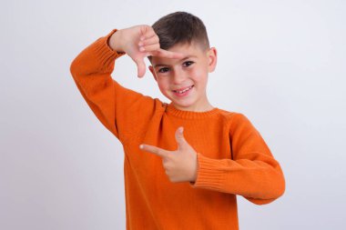 Beyaz arka planda duran örgü turuncu süveter giyen beyaz çocuk elleriyle parmak çerçevesi yapıyor. Yaratıcılık ve fotoğrafçılık kavramı.
