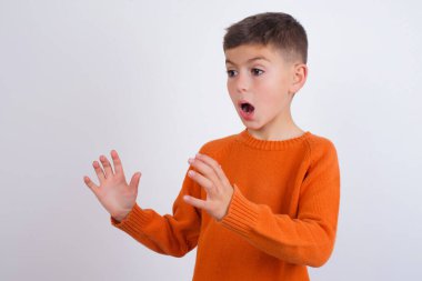 Beyaz arka planda duran örgü turuncu süveter giyen beyaz çocuk yüksek sesle bağırıyor, gözleri açık ve elleri gergin.