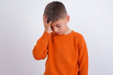 Beyaz arka planda dikilip ağlayan, örgü örmüş turuncu kazak giyen çok üzgün ve yalnız bir beyaz çocuk.,