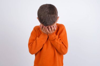 Kafkas çocuk örgü turuncu süveter giyiyor beyaz arka planda duruyor yüzünü elleriyle kapatıyor, yıkılmış ve ağlıyor. Üzücü bir kavram