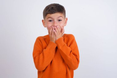 Beyaz arka planda dikilen örgü turuncu süveter giyen neşeli beyaz çocuk, neşeyle kıkırdıyor, ağzını kapatıyor, doğal kahkahaları var, olumlu hikayeler duyuyor ya da komik anekdotlar dinliyor.