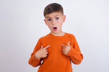 Beyaz arka planda dikilen örgü turuncu süveter giyen utangaç beyaz çocuk, yarışmaya katılmak için seçildiği için şaşkın bir ifadeyle, bir konuda tereddüt içinde olduğunu gösteriyor.