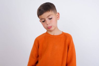 Kasvetli, reddedilmiş, beyaz arka planda duran örgü turuncu süveter giyen beyaz çocuk sorunlar ve zorluklar yaşıyor, alt dudak kıvrımları var ve umutsuzluk içinde gözlerini kapatıyor, depresyonda.