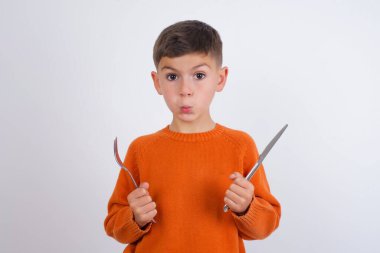 Örgü örmüş turuncu süveter giyen beyaz arka planda duran, çatal bıçaklı, manken gibi aç beyaz çocuk lezzetli bir pizza turtası istiyor.