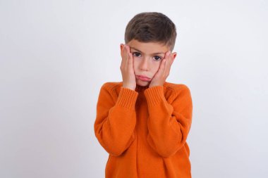 Beyaz arka planda örülü turuncu süveter giyen beyaz çocuk. Yüzü, depresyonu ve üzüntüyü örten yorgun eller.