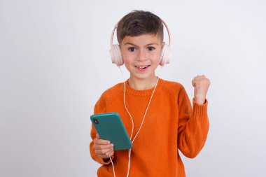 Beyaz arka planda dikilen örgü turuncu süveter giyen beyaz çocuk, kulaklıklara bağlı modern cep telefonunu tutuyor, iyi duygulardan yumruğunu sıkıyor, sevinçle haykırıyor.,