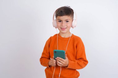 Beyaz arka planda duran örgü turuncu süveter giyen mutlu beyaz çocuk akıllı telefon ekranına odaklanırken iyi hissediyor. İnsanlar, teknoloji, yaşam tarzı