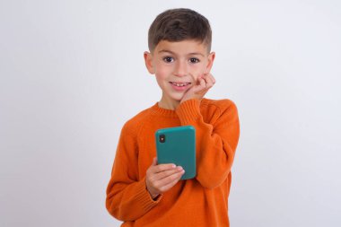 Beyaz arka planda dikilen örgü turuncu süveter giyen neşeli beyaz çocuk cep telefonu cihazıyla poz veriyor, modern akıllı telefona mesaj yazıyor, boş zamanlarında komik videolar izliyor, iyi internet bağlantılarından hoşlanıyor.,