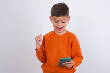 Beyaz arka plan üzerinde örgü örmüş turuncu süveter giyen neşeli beyaz çocuk hücrede el ele kitap okuyor.