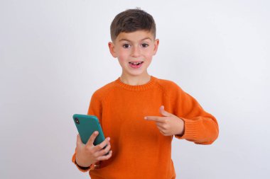 Beyaz arka planda dikilen örgü turuncu süveter giyen beyaz çocuk yeni bir hücre gösteriyor.,