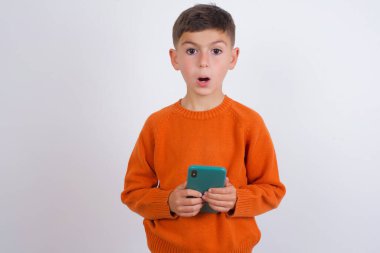 Beyaz arka planda dikilen örgü turuncu süveter giyen Kafkasyalı çocuk ağzı açık telefon reklamı okuyor inanılmaz yüksek alışveriş fiyatları.