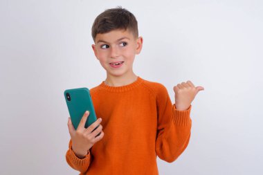 Beyaz arka plan üzerinde örülü turuncu süveter giyen beyaz çocuk akıllı telefonuyla mesaj atıyor ve gülümseyen bir gülümsemeyle baş parmağıyla onu işaret ediyor.